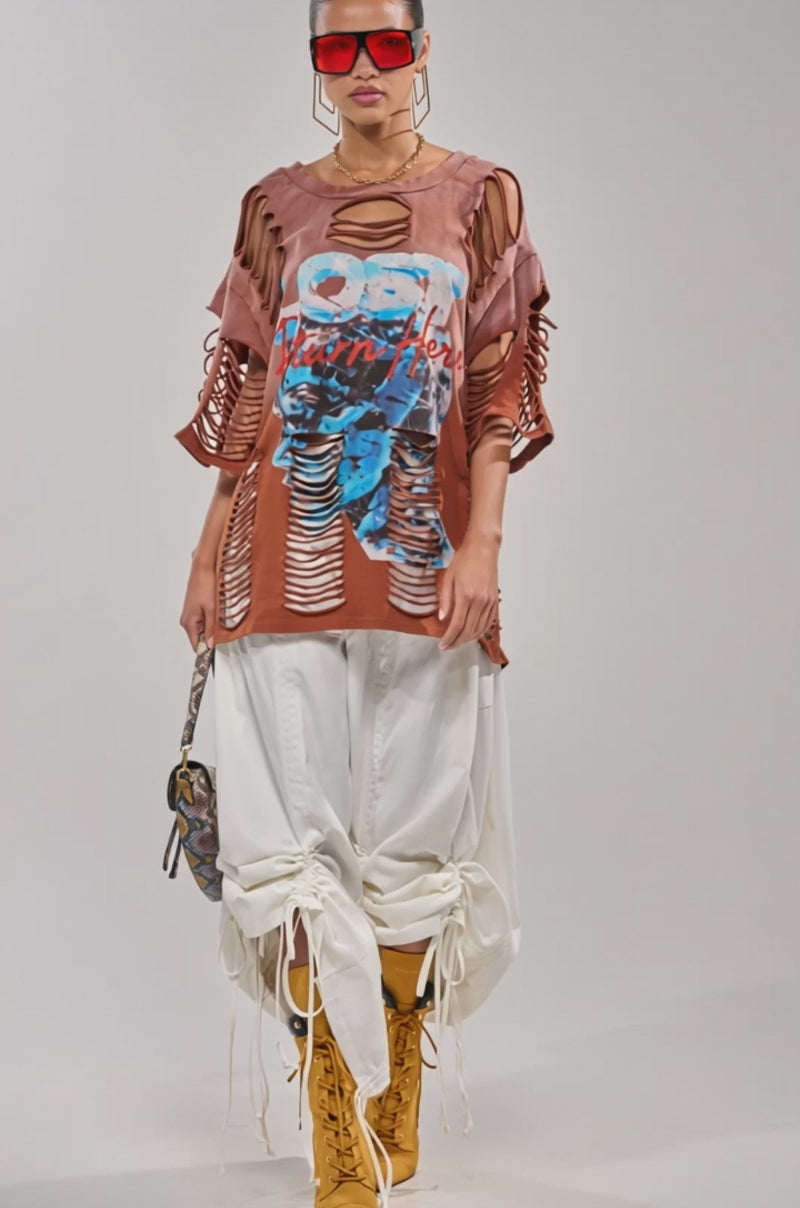 A video of a woman wearing oversized red sunglasses, the BYE GIRL OVERSIZED DISTRESSED TEE IN BROWN, loose white pants tied at the ankles, tan lace-up boots, gold hoop earrings, and carries a small patterned bag.