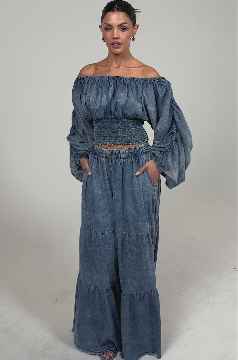 Video of woman modeling the DENIM FOR DAYS PALAZZO PANT. Denim-hued, tiered palazzo pants with an off-the-shoulder voluminous-sleeved top, hoop earrings, and sandals, with her hair pulled back.