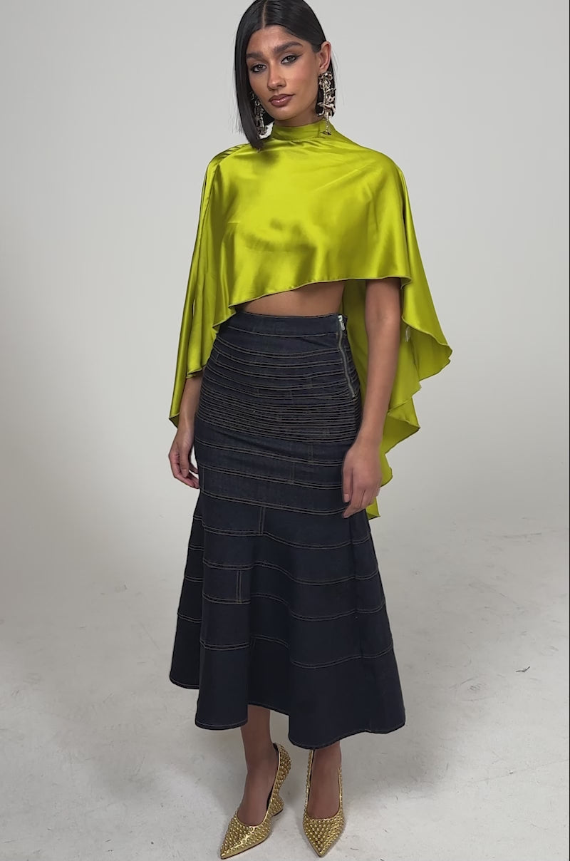 A video of a woman in the BABY MAKE A MOVE BLOUSE IN CHARTREUSE, featuring shiny satin fabric and draped sleeves, paired with a high-waisted dark denim mermaid skirt, gold glitter heels, straight dark hair, and statement earrings.