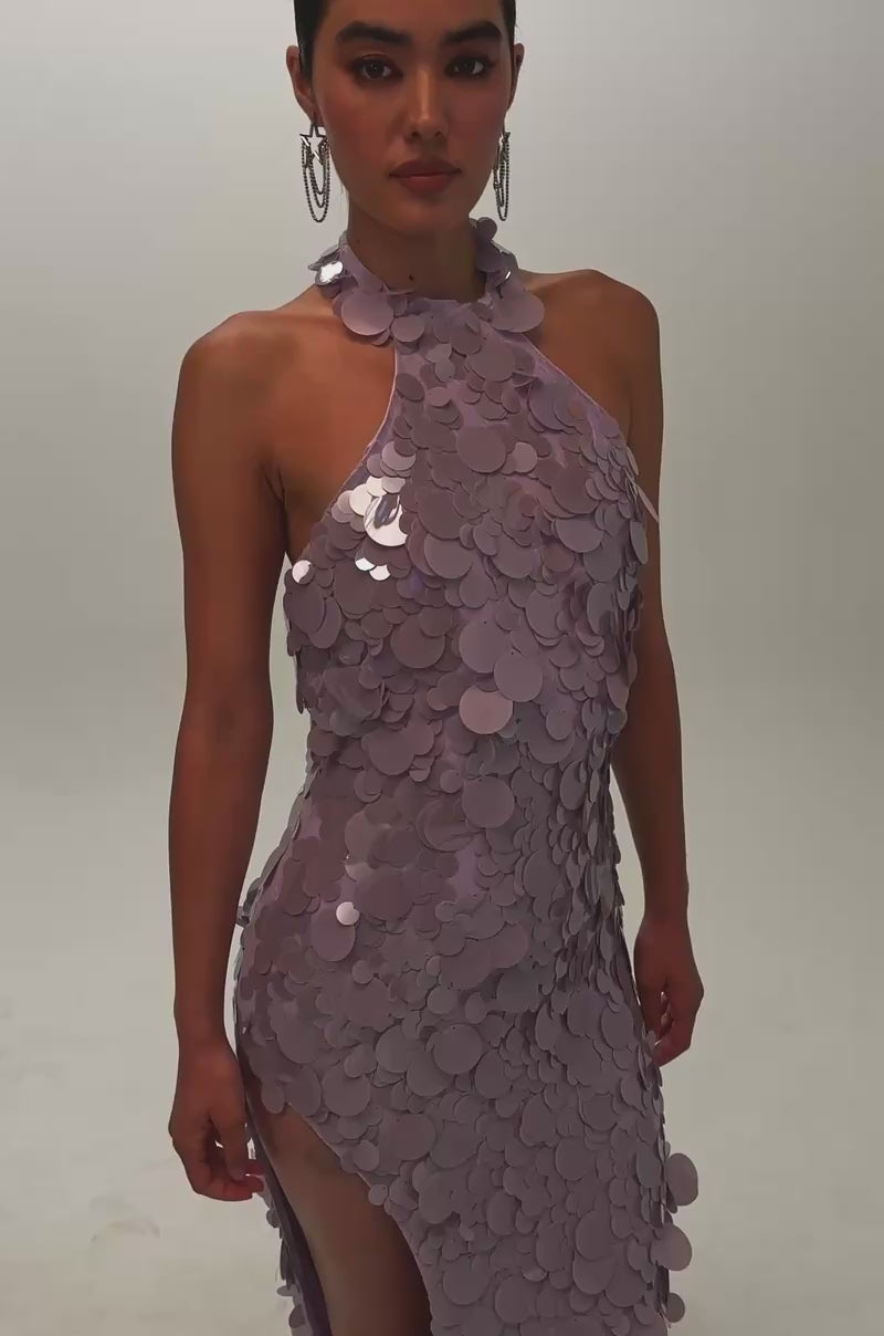 Video of a woman modeling the FALL FOR YOU SEQUIN MAXI DRESS IN LILAC—a sleeveless, mock-neck maxi dress embellished with large lilac sequins, featuring a thigh-high slit.