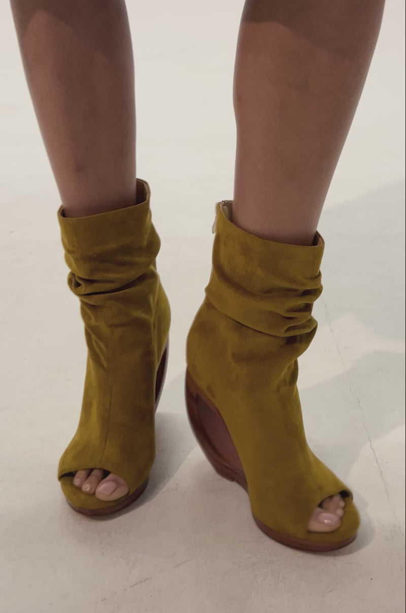 Video of woman modeling the AZALEA WANG ARLO Mustard Wedge Open Toe Faux Suede Bootie, featuring curved wooden wedge heels, standing on a light background.