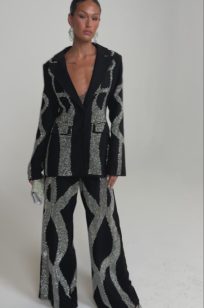 A video of a woman in the GEMINI RHINESTONE PANT, a black high-rise, wide-leg trouser adorned with bold silver crystal patterns, paired with a matching blazer, large hoop earrings, and a clutch.