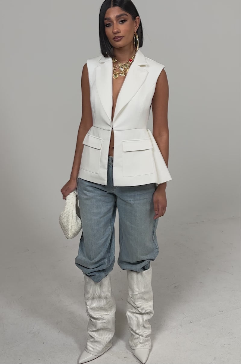 A video of a woman wearing the NAOMI BLAZER VEST IN WHITE styled with light blue baggy jeans, tall white textured boots, a white woven clutch, and layered gold necklaces.