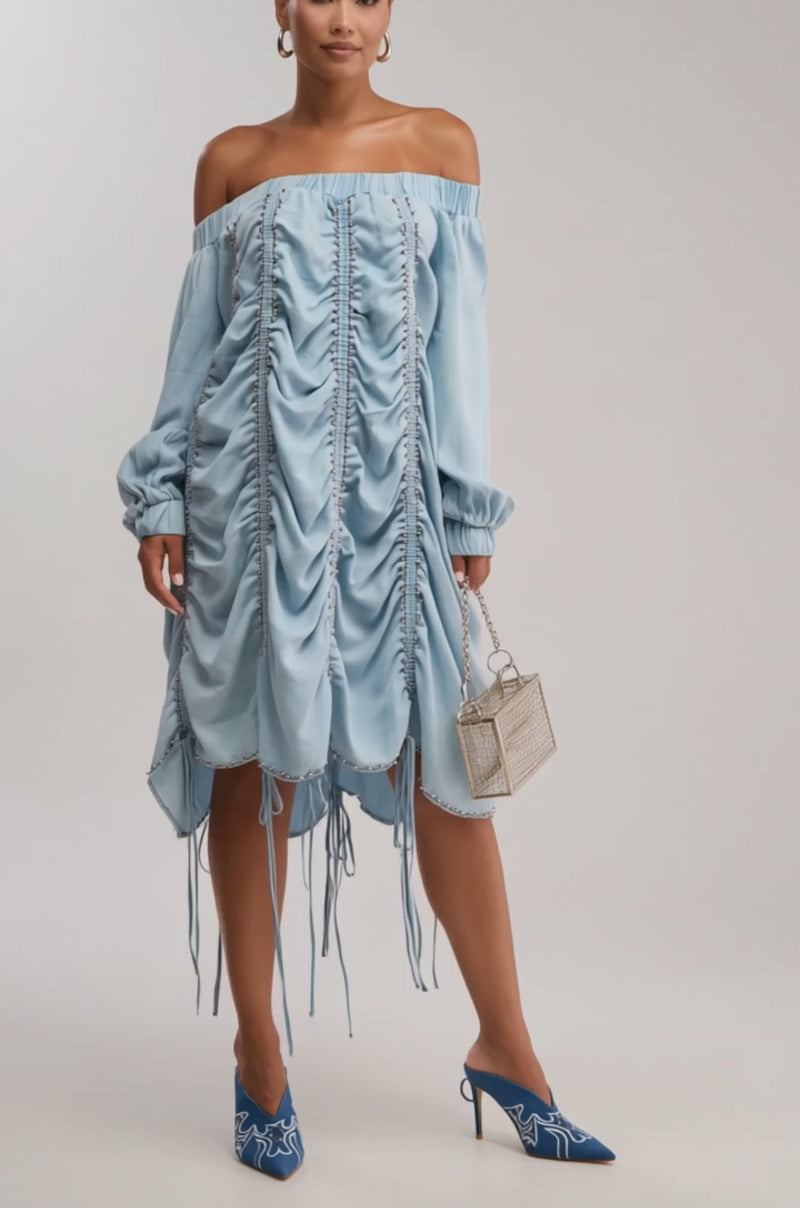Video of woman modeling the FREE AS CAN BE MINI DRESS IN LIGHT DENIM, featuring off-the-shoulder ruched details and ties, blue heels, hoop earrings, and a small cage-shaped purse—effortlessly embodying chic denim style.