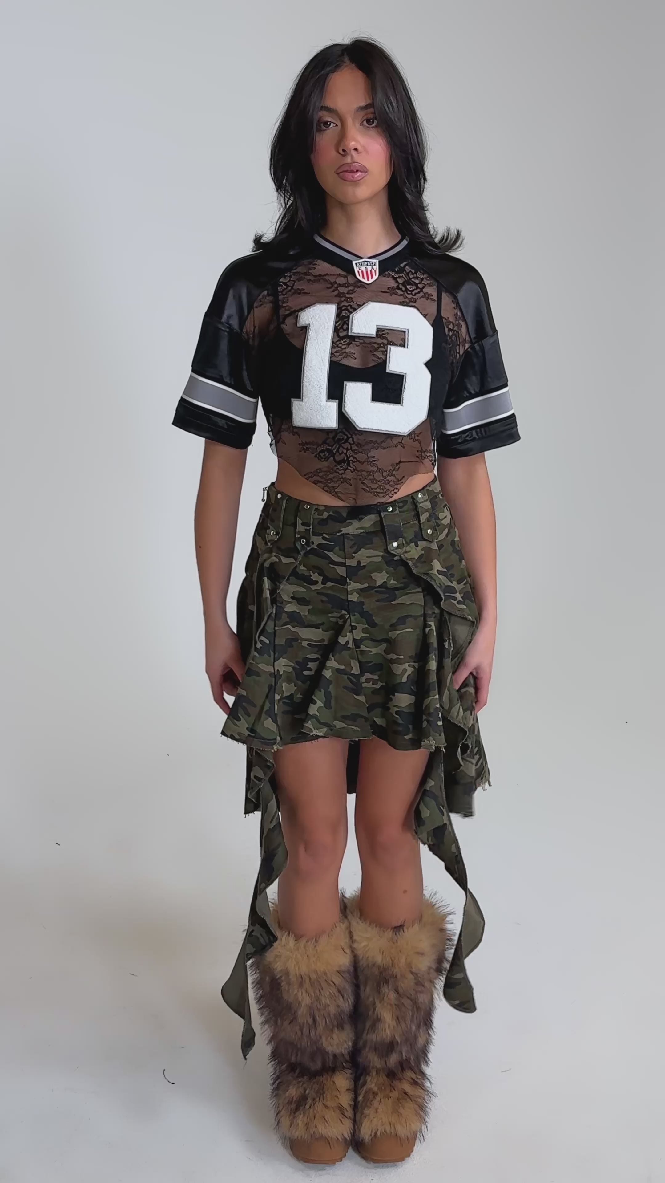 A video of a woman wearing a black mesh football jersey with the number 13, the IN THE TRENCHES CAMO MINI SKIRT with ties, and furry brown boots. 