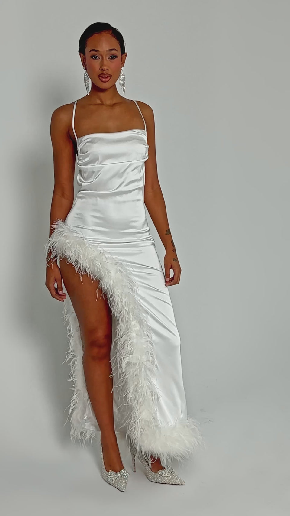 Video of woman modeling the LIGHT AS A FEATHER MAXI DRESS, featuring a white feather trim, thin straps, and a high thigh slit. She pairs it with sparkly heels and statement earrings against a plain light background.