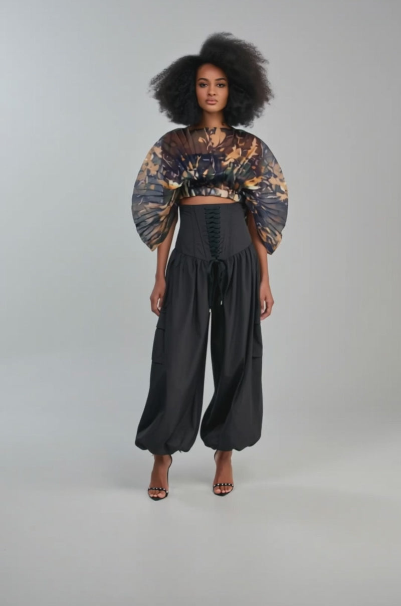 Video of a woman modeling the MIDNIGHT MIRAGE CAMO TOP, featuring wide, flowing pleated sleeves, styled with a fitted black corset.
