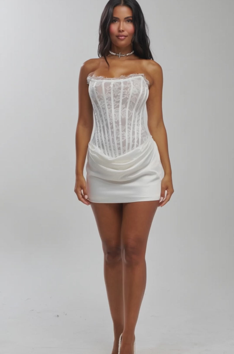 Video of woman modeling the SPOIL ME LACE MINI DRESS IN WHITE, featuring a lace bodice, satin mini skirt, and elegant draping. She pairs it with a pearl choker and wears her long dark wavy hair against a plain light gray background.