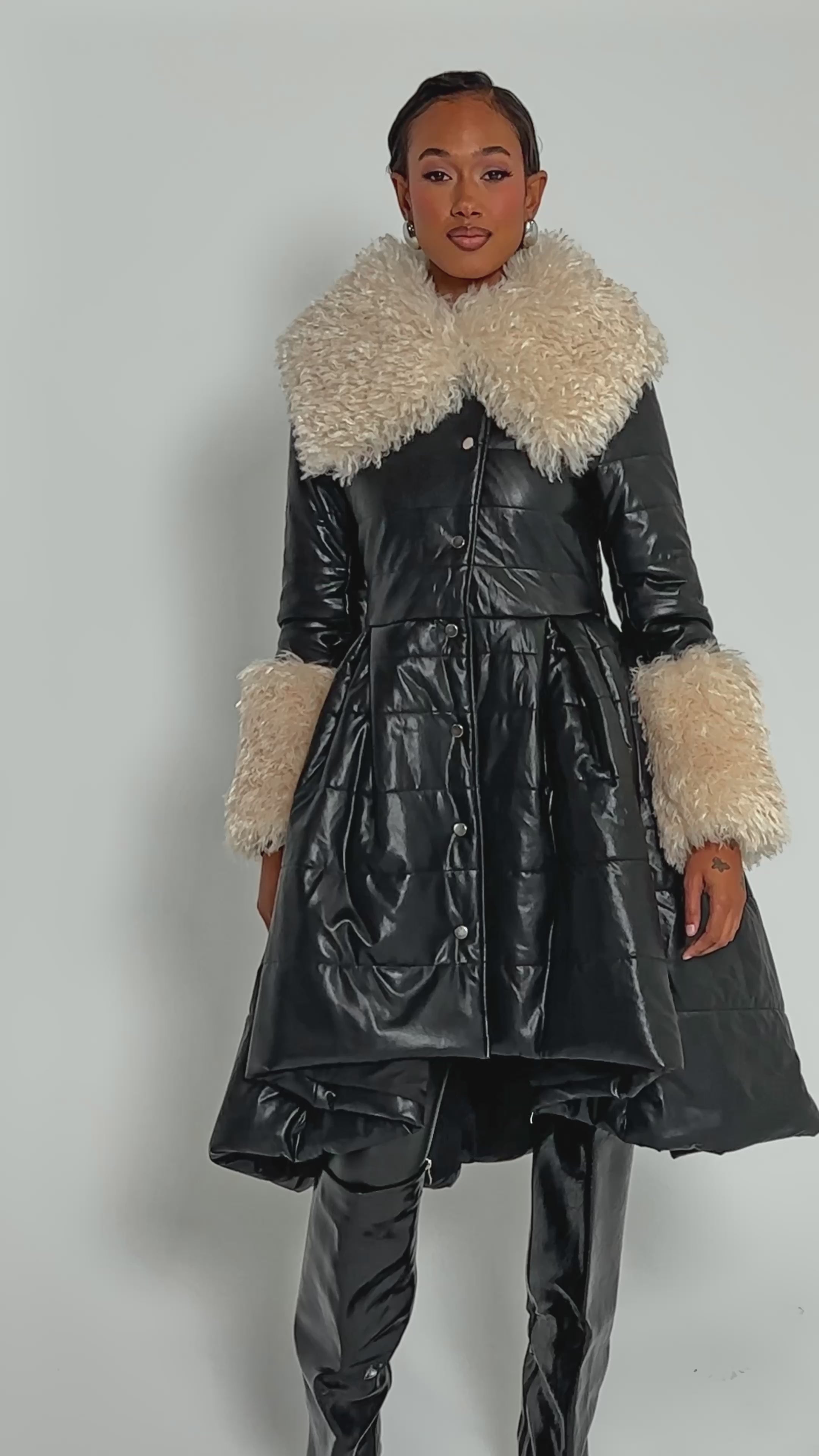 Video of woman modeling the MIDTOWN PEPLUM PUFFER IN FAUX LEATHER—a black, fitted jacket with large cream faux fur trim on the collar and cuffs.