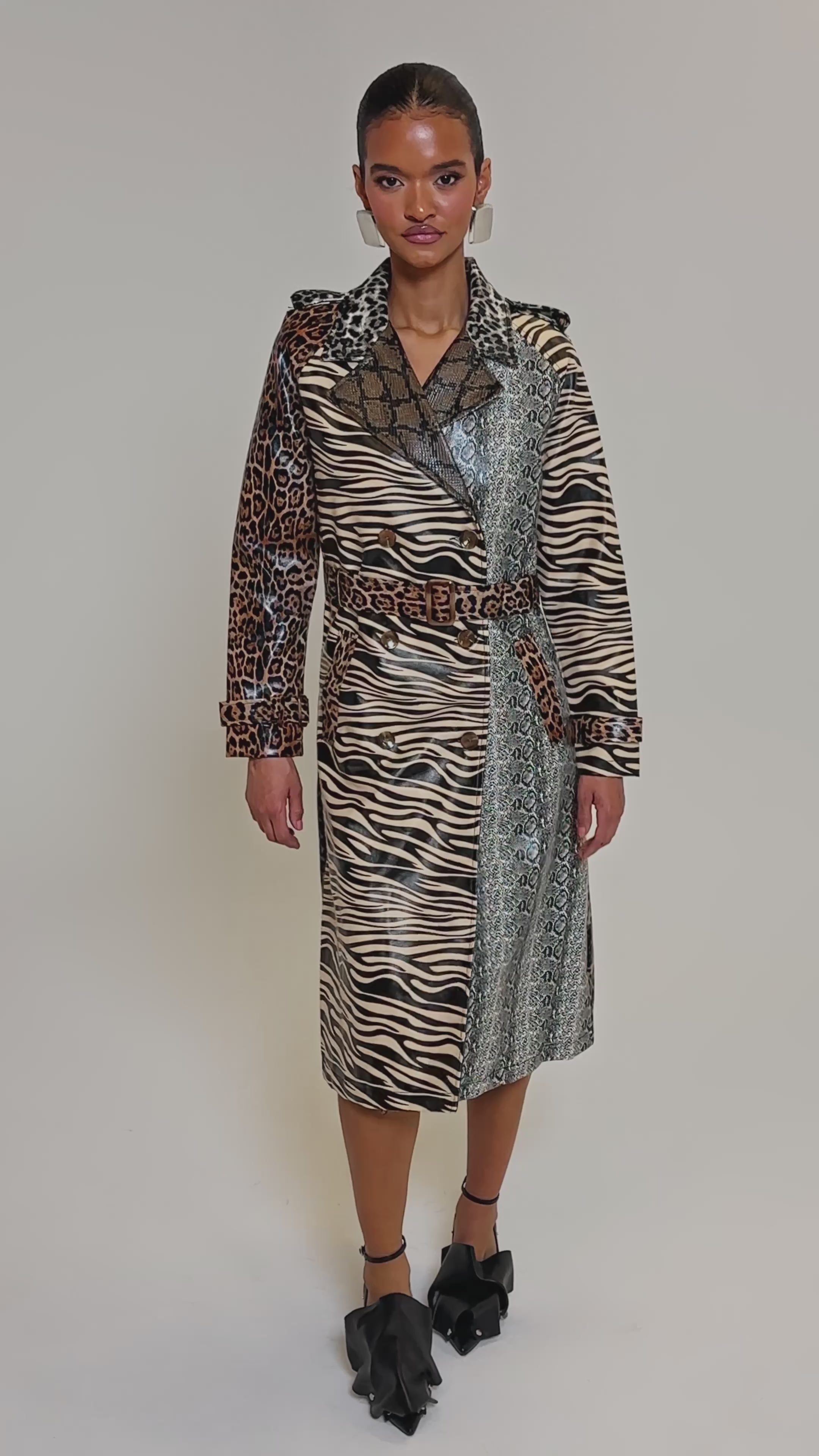 Video of woman modeling the BASEL PAINTED TRENCH, a faux leather coat with zebra, leopard, and snakeskin prints. She pairs it with black high-heeled sandals featuring bows and wears her hair slicked back against a plain backdrop.