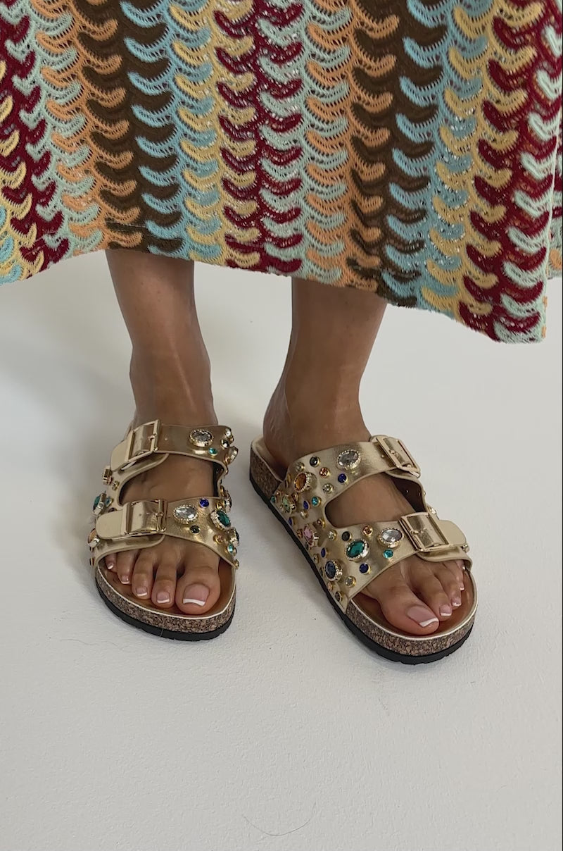 A video of a person wearing a colorful patterned skirt and the AZALEA WANG BAYLEAVES GOLD EMBELLISHED SANDAL, featuring multicolored gems and metallic buckles.