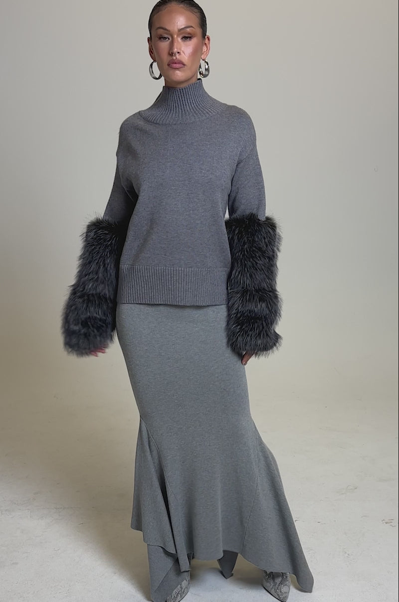 A video of a woman posing in the TOUCH ME SWEATER WITH FUR CUFFS, a light gray turtleneck with dark gray fur cuffs, paired with a matching long skirt with a flared asymmetric hem, set against a plain light gray background.