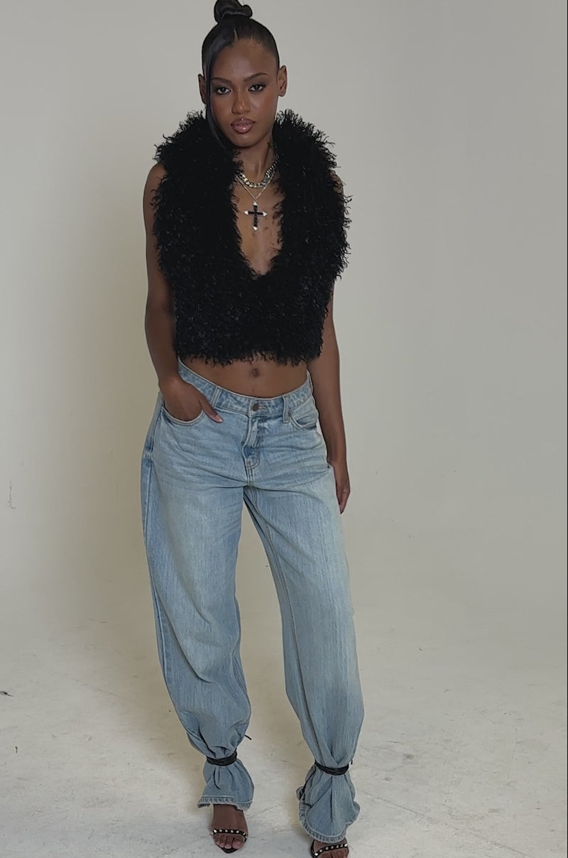 Video of woman modeling the THINK ABOUT YOU ALL THE TIME FUR BLOUSE IN BLACK, a sleeveless, hooded, deep V-neck top, paired with light blue jeans, layered necklaces, and a sleek updo with side-swept bangs against a plain background.