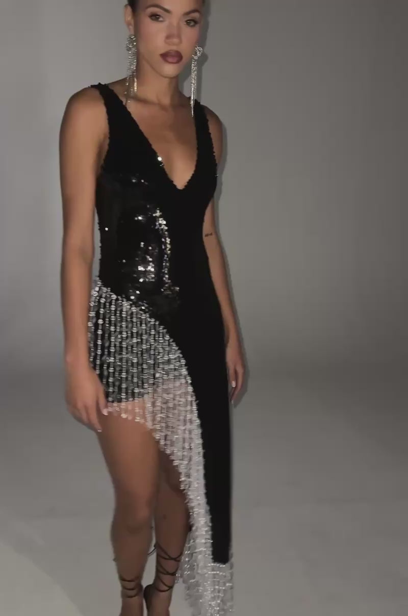 Video of woman modeling the NEW MONEY SEQUIN BLOUSE, featuring sleeveless black sequins, an asymmetrical hem, and fringe beaded trim, styled with strappy black heels and statement earrings.