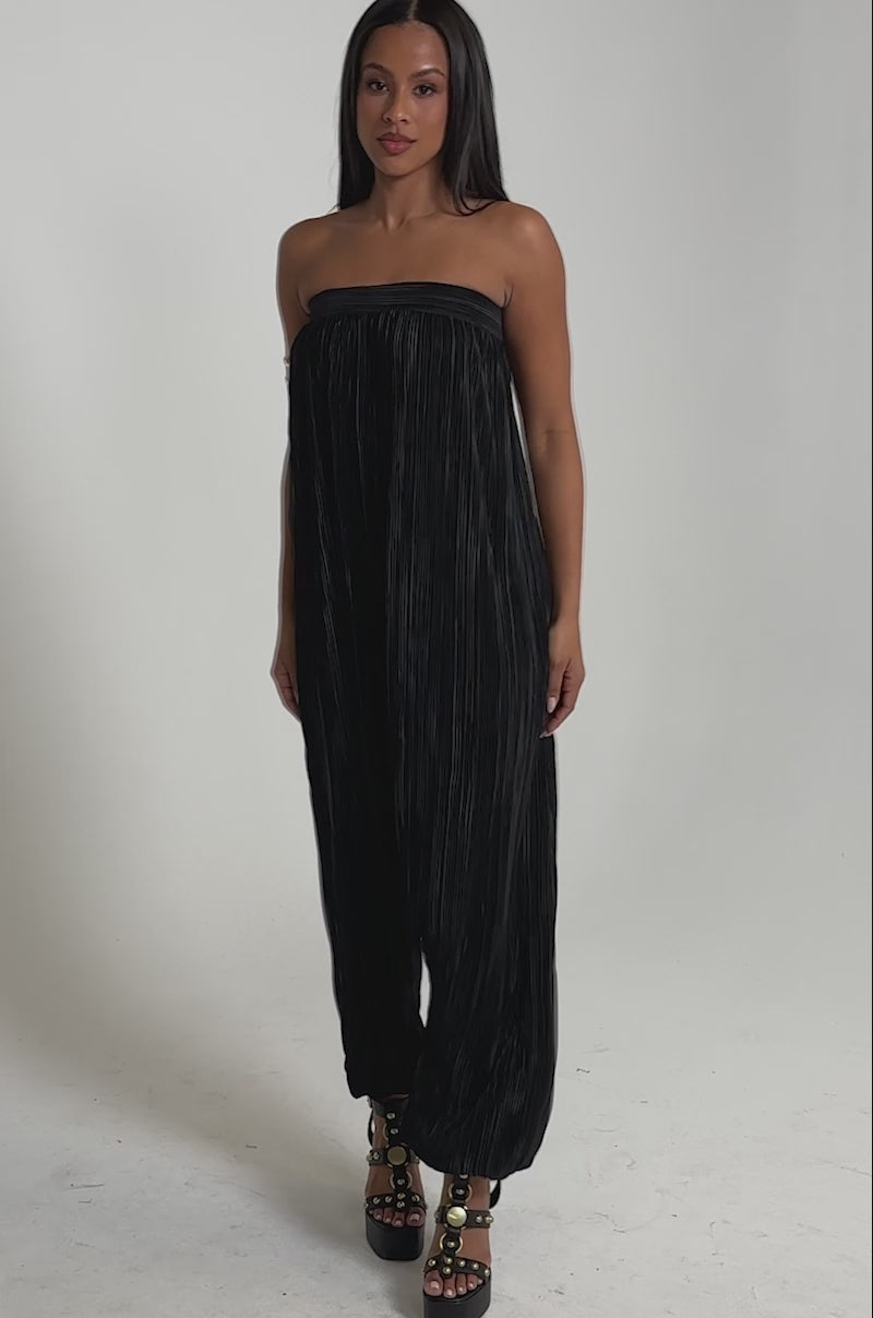 A video of a woman in the FROM THE CITY DROP CROTCH JUMPSUIT IN BLACK, a loose, pleated, strapless evening resort style. 