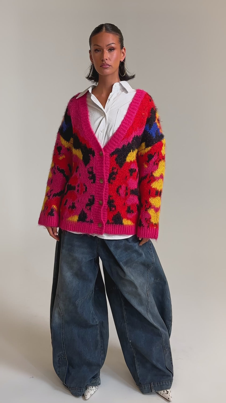 A video of a woman wearing the COZY VIBES CARDIGAN, an oversized, multi-color jacquard piece with a bold pattern, styled over a white collared shirt and wide-leg blue jeans against a plain light background.