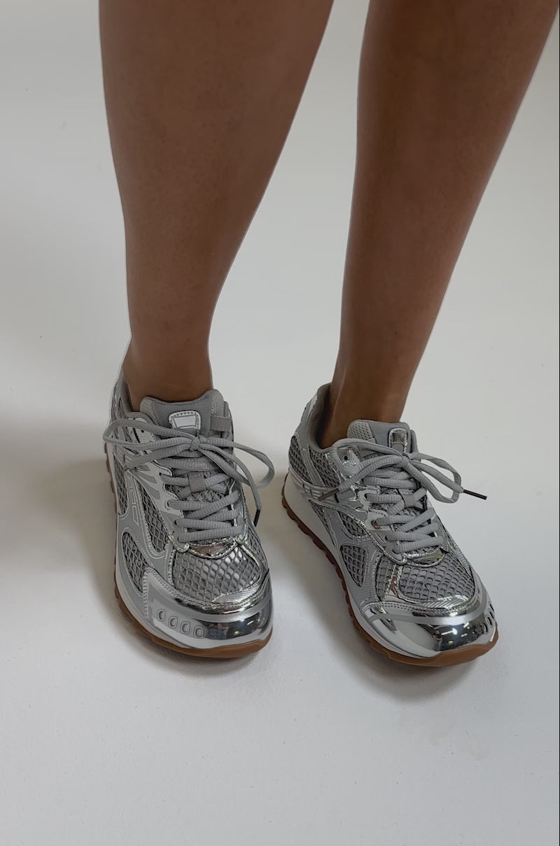 A video of a person wearing the AKIRA GOLD LENIYA SILVER SNEAKER, featuring metallic silver with mesh details, light gray laces, and brown rubber soles.