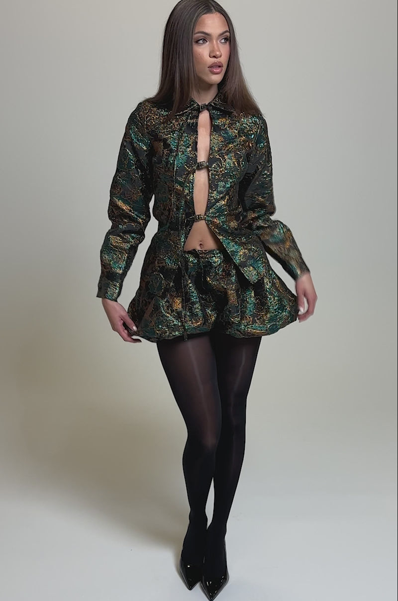 Video of woman modeling the TOO HOT TO HANDLE BLOUSE featuring a metallic brocade finish and keyhole cutout, paired with a voluminous skirt and dark tights against a plain background.