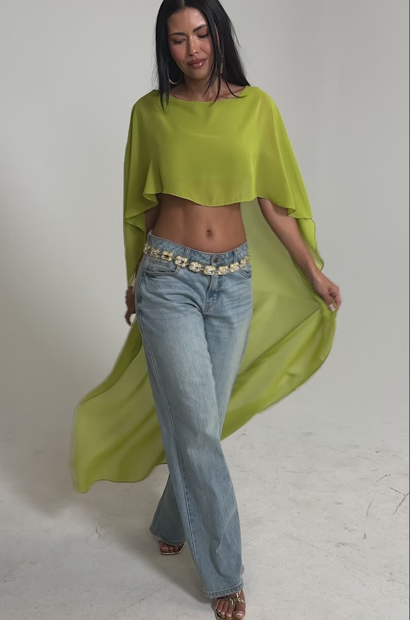 Video of woman modeling the IT GIRL BLOUSE IN CHARTREUSE—a flowing chiffon cape top—styled with light blue wide-leg jeans, a decorative belt, and sandals for chic resort wear against a plain white background.