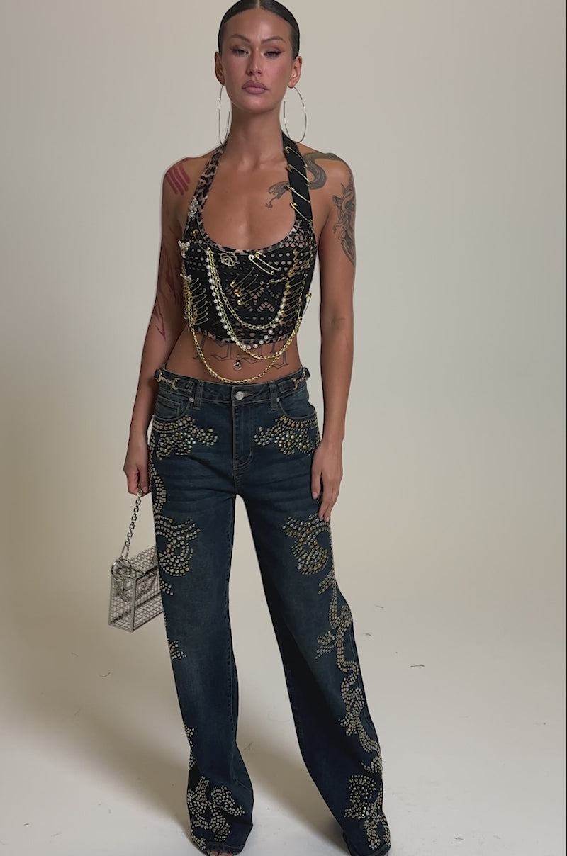 A video of a woman in the ZIGGY STUDDED WIDE LEG DENIM, paired with a beaded crop top and a metallic bag, her arm tattoos add edge to the bold, gold-studded look.