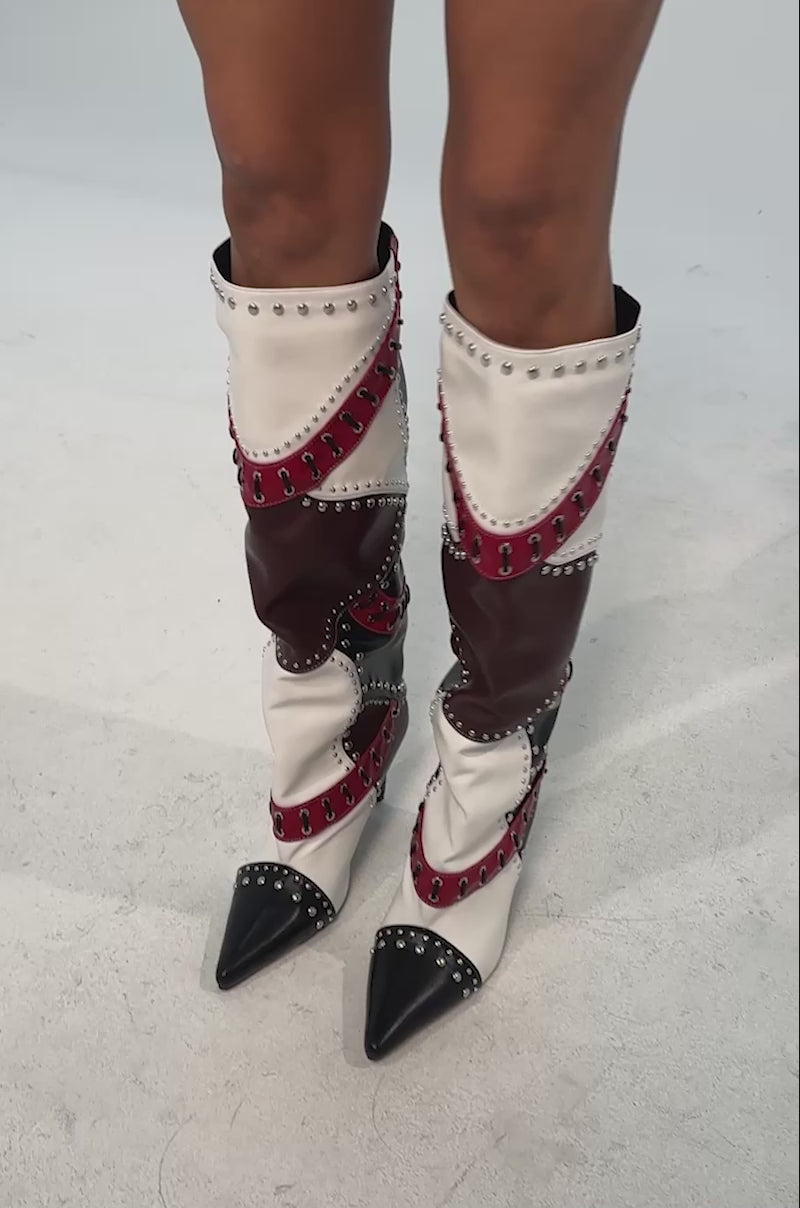 Video of woman modeling the AKIRA GOLD VESSAE PATCHWORK STILETTO BOOT, a knee-high leather boot with red, white, black, and maroon panels, silver studs, lace-up details, a pointed toe, and a stiletto heel.