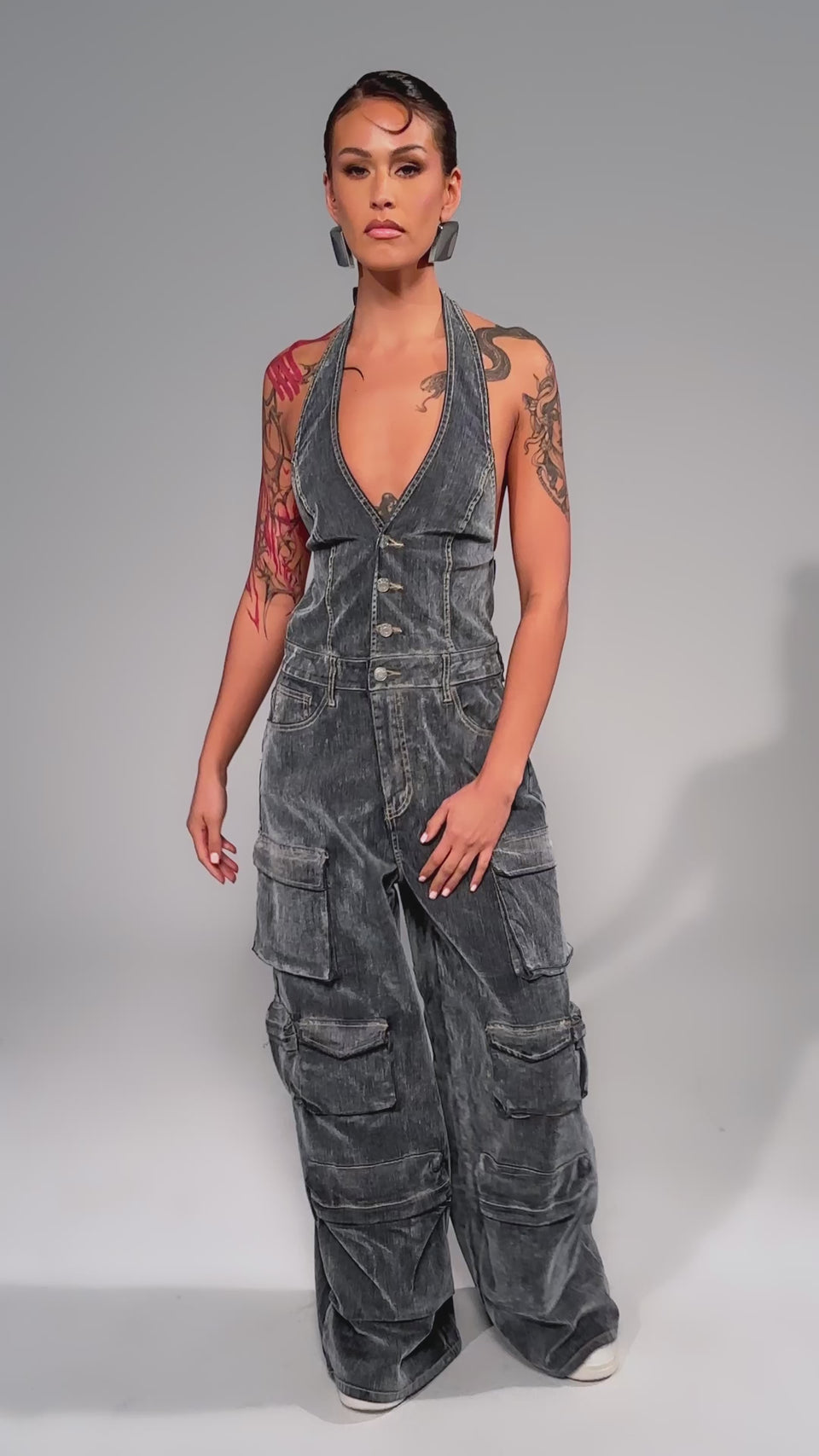 Video of woman modeling the MELINDA VELVET JUMPSUIT—sleeveless, deep V-neck, wide-leg style with multiple cargo pockets—paired with hoop earrings, white sneakers, and arm tattoos visible.