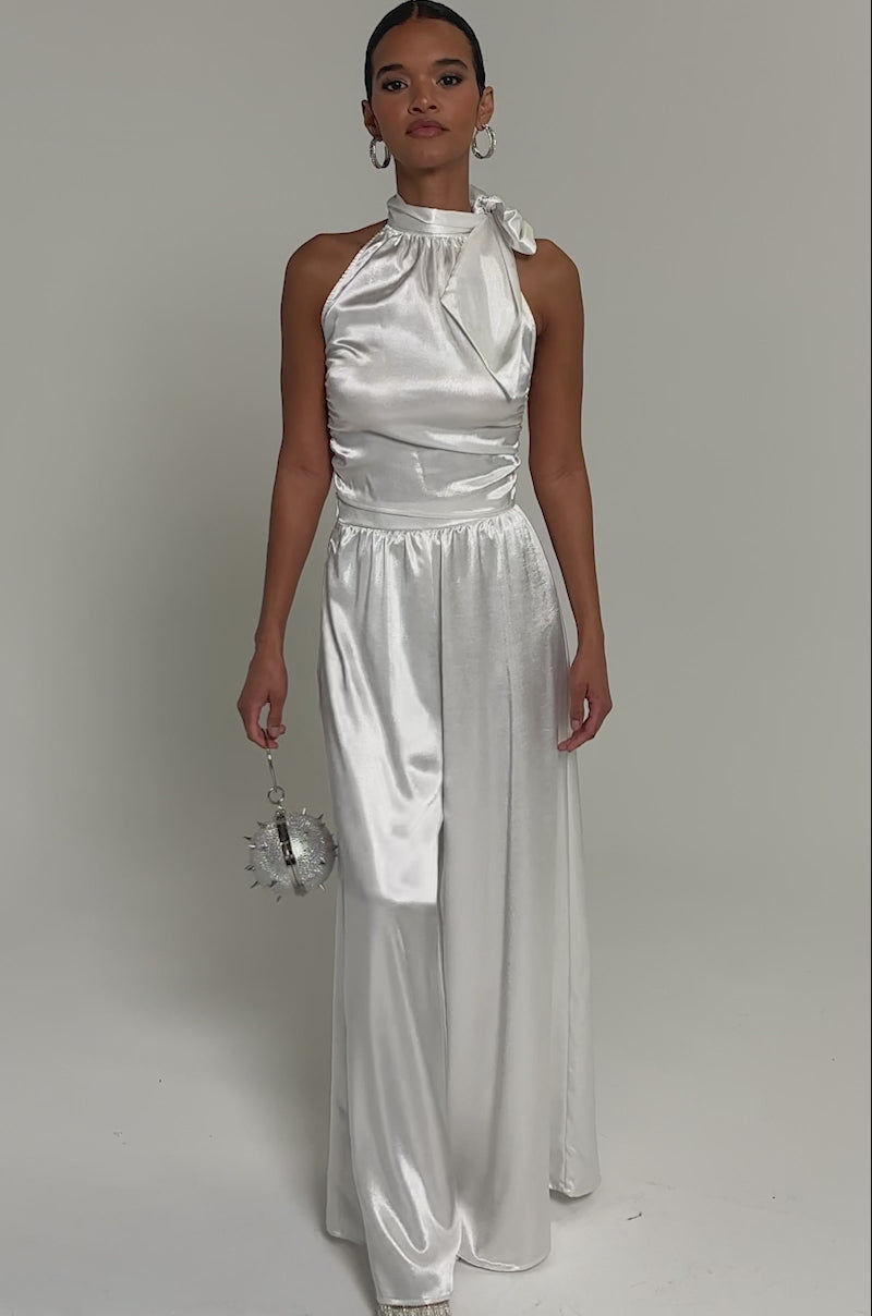 Video of woman modeling the LEAVE ME ALONE SLINKY PALAZZO PANT, a sleeveless silky white high-rise wide-leg pants. She holds a spiked purse and accessorizes with pointed shoes and hoop earrings.