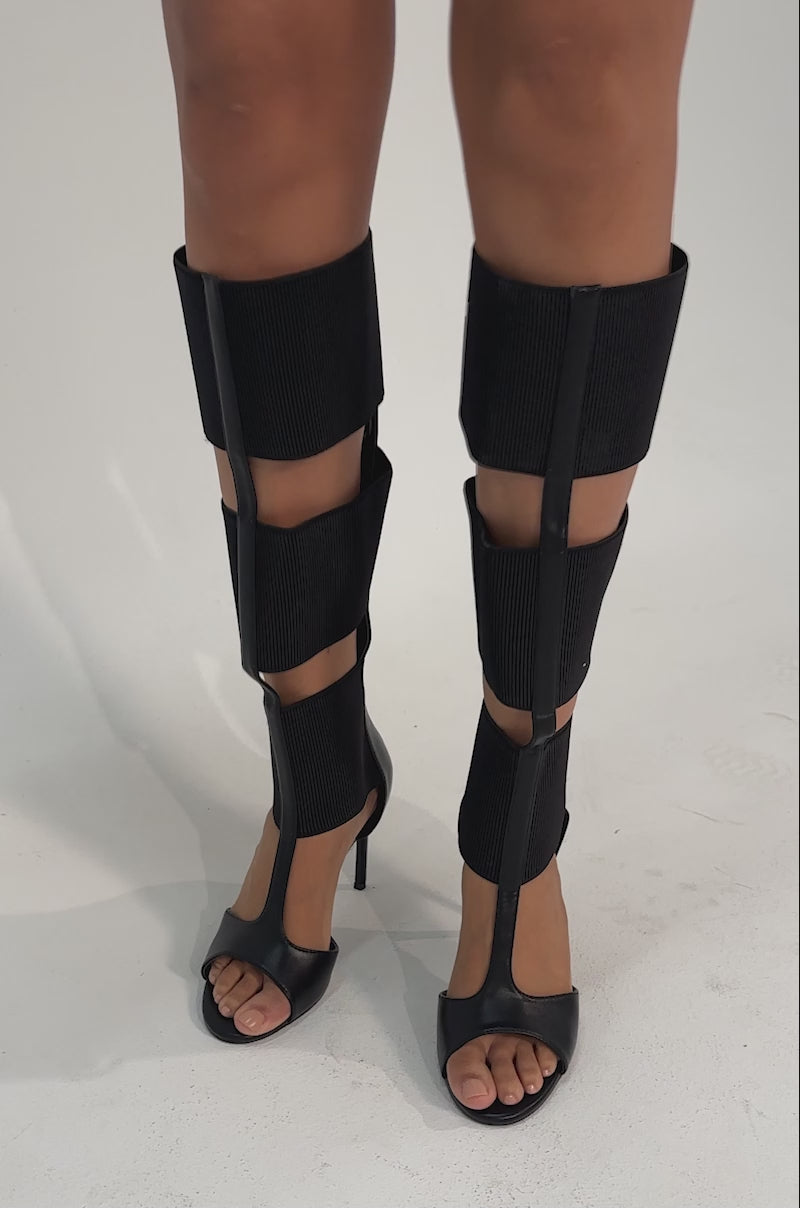 A video of a person wearing AKIRA GOLD BELLANTRA BLACK GLADIATOR STILETTO SANDALS with unique black leg wraps, revealing their knees and lower thighs.