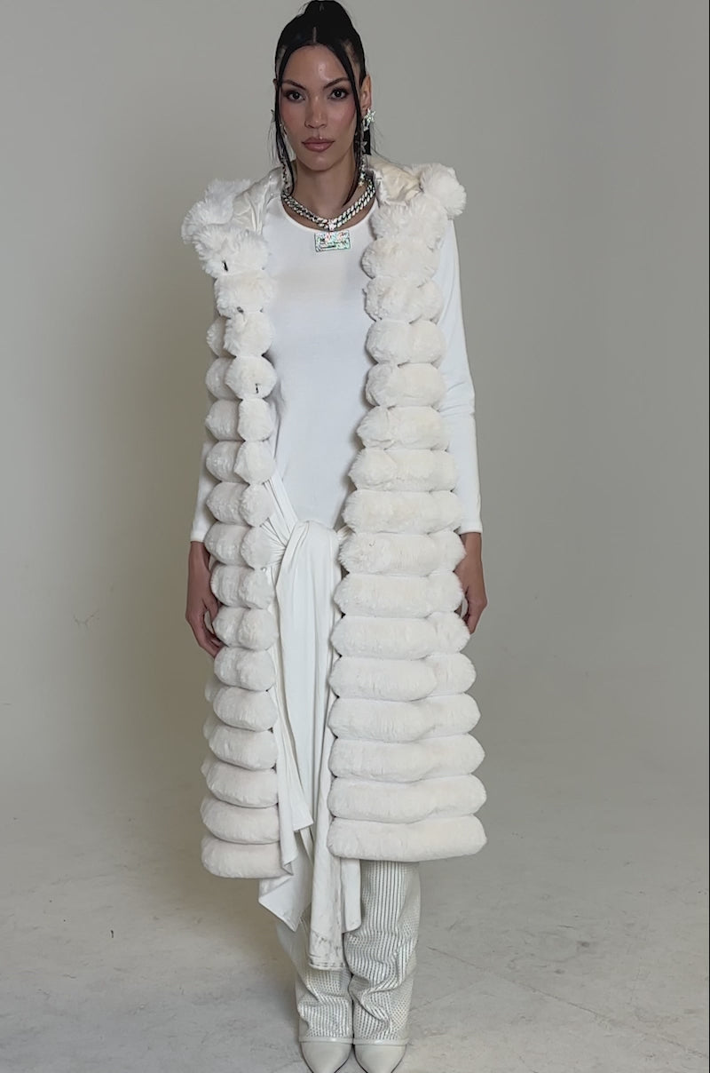 A video of a woman posing in the POPPY FAUX FUR HOODED VEST, styled with wide-leg pants and a knotted accent. She finishes her chic winter look with high heels, jewelry, and slicked-back hair for a sophisticated ensemble.