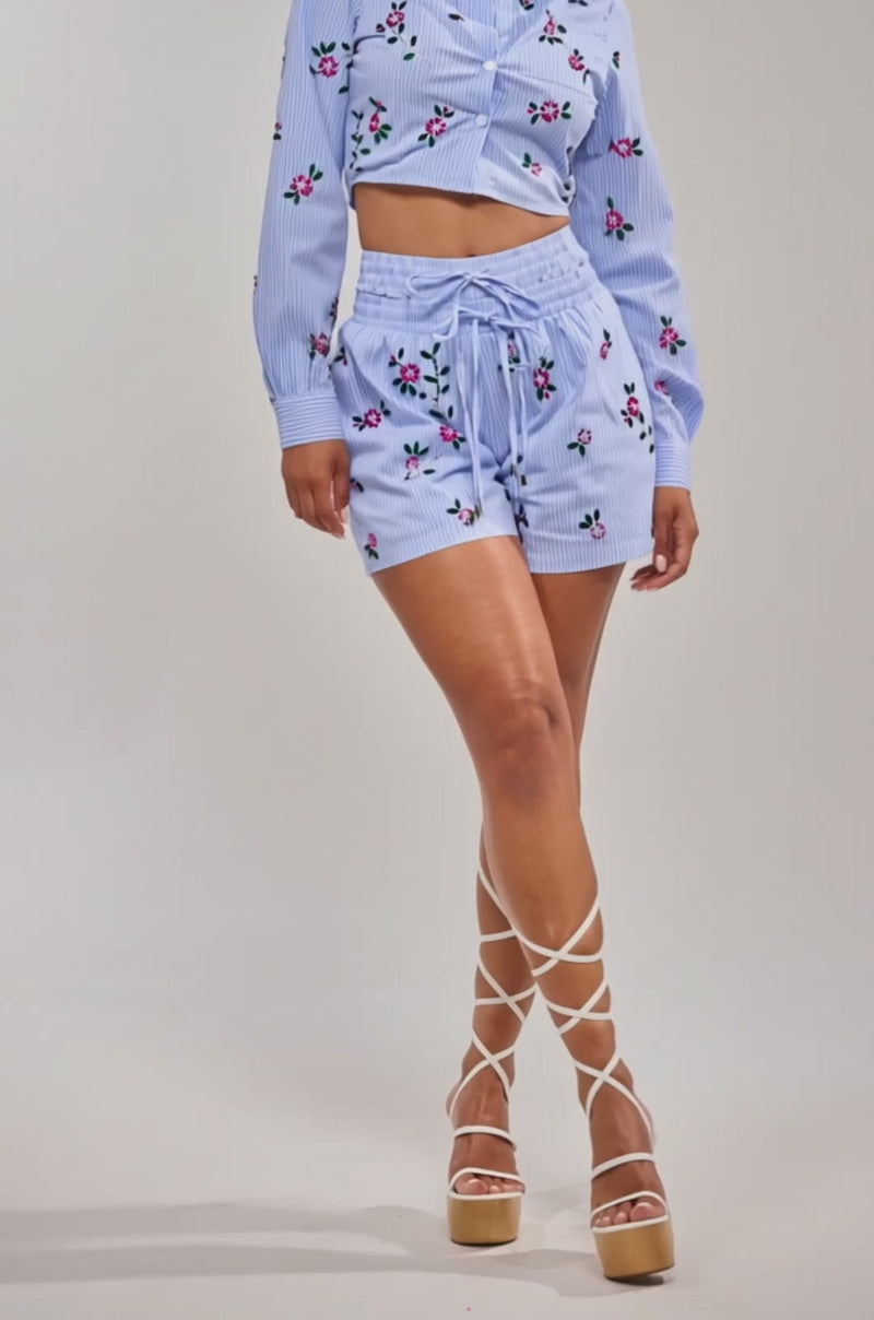 A video of a woman in the JUNIE POPLIN RHINESTONE DETAIL SHORT, a light blue floral print statement short paired with a matching collared crop top and lace-up platform sandals. 