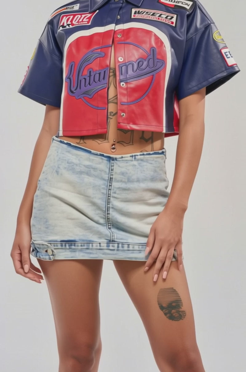 Video of woman modeling the PEDAL TO THE METAL FAUX LEATHER BUTTON DOWN featuring "Untamed" on the front, paired with a light denim skirt, hoop earrings, slicked-back hair, and a torso tattoo—an ideal streetwear look for festivals.