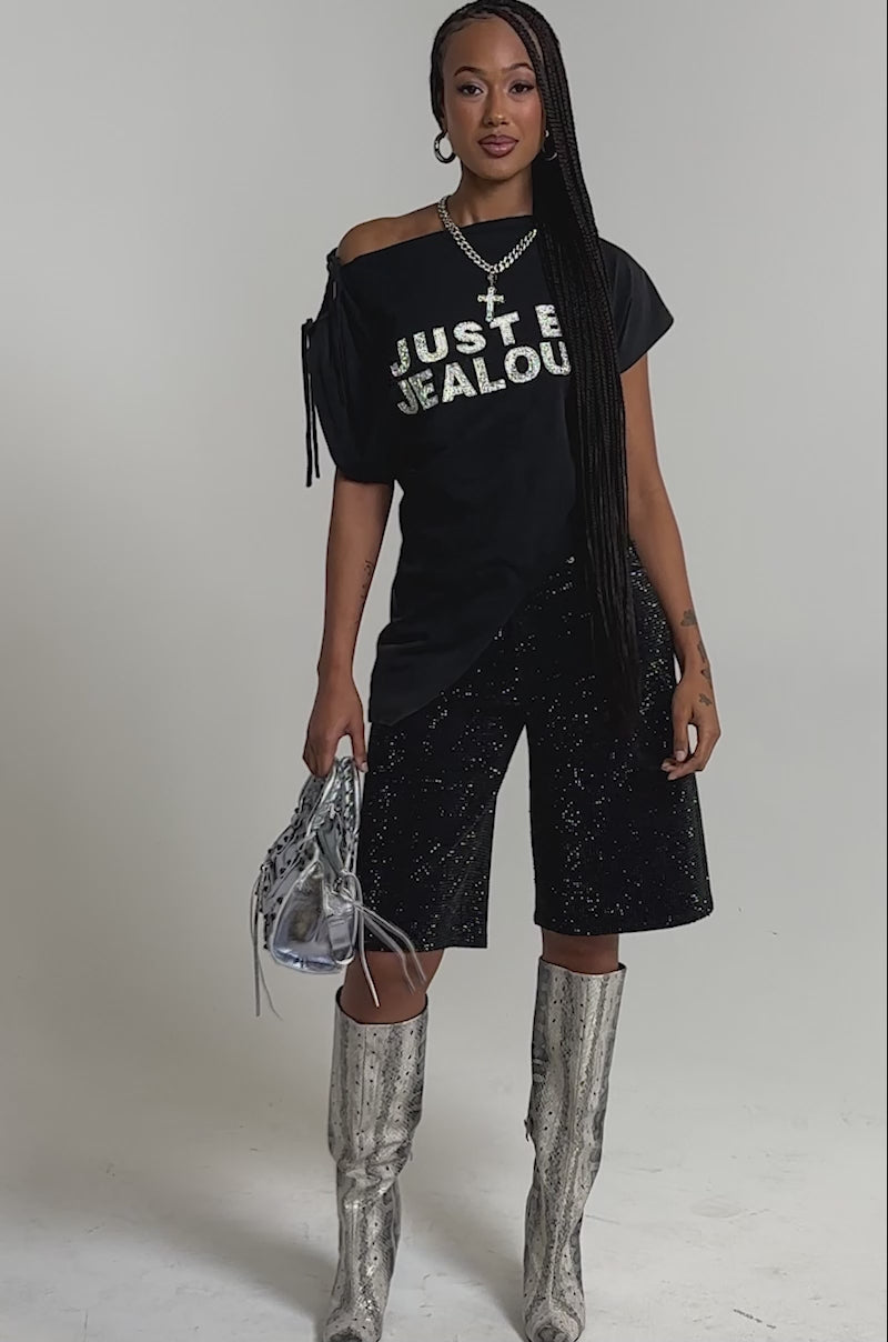 A video of a woman wearing a black “JUST BE JEALOUS” t-shirt, BLING BLING RHINESTONE BERMUDA SHORT, silver knee-high boots, layered necklaces, and holding a shiny silver handbag.