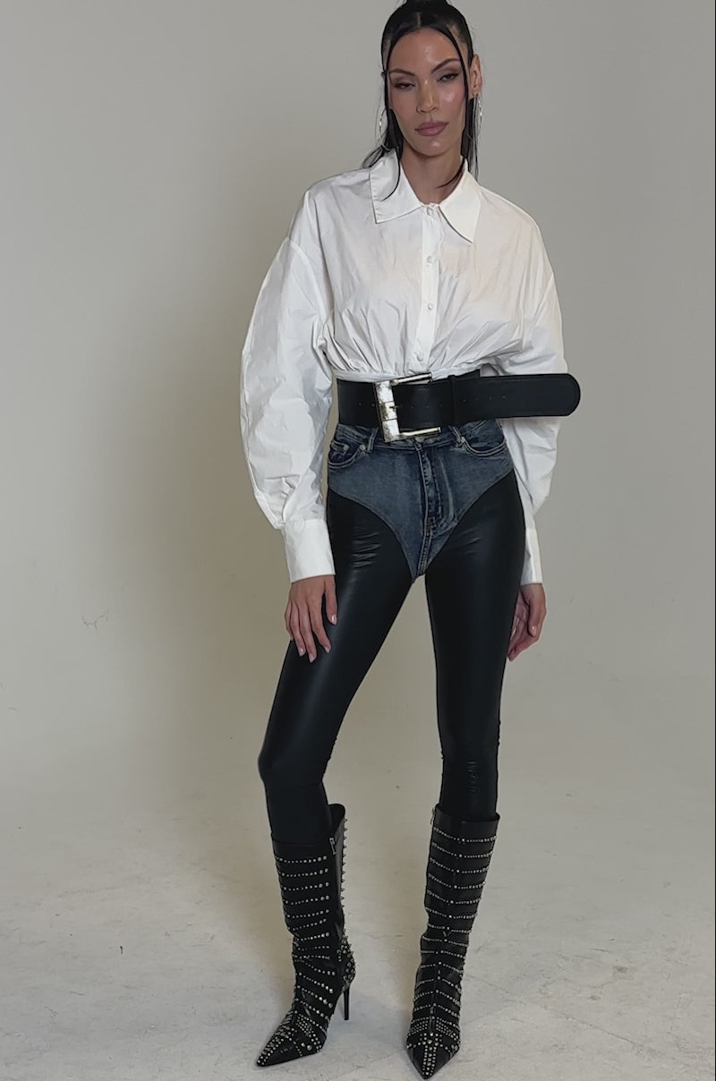 A video of a woman posing in the LET'S GO BABY BODYSUIT, high-waisted denim, black leather-look pants, a faux leather belt, and knee-high studded black boots. Her long dark hair is styled in a high ponytail.