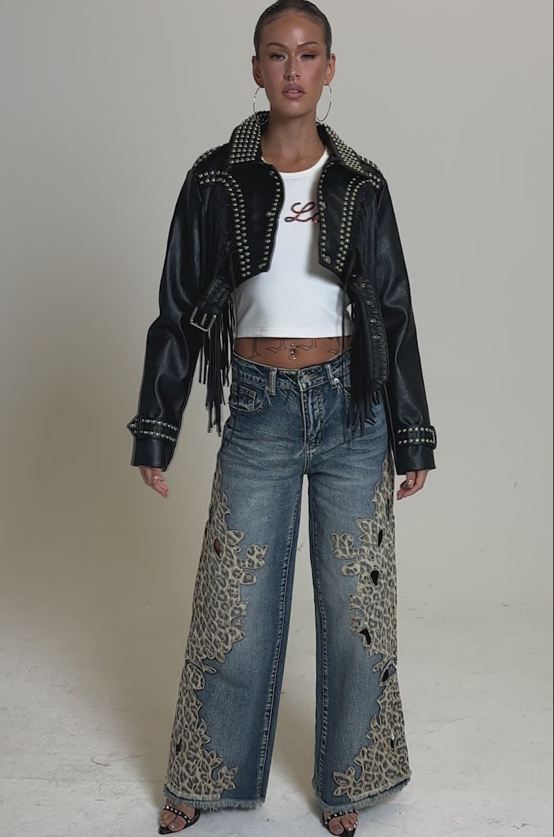A video of a woman posing in cheetah print wide leg pants, a white crop top, studded black leather jacket with fringe, heeled sandals, against a plain background.
