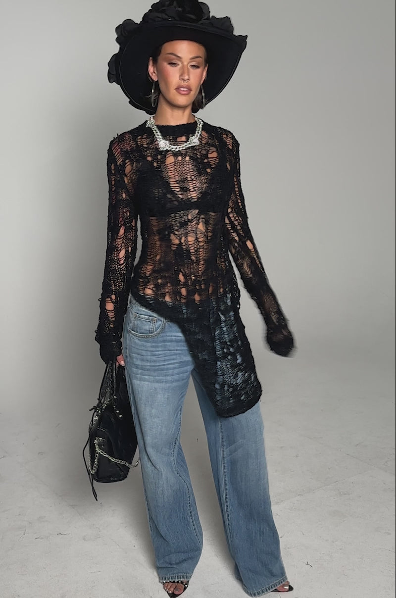 A video of the ASYMMETRICAL KNIT SWEATER over a black bra, pairing it with wide-leg blue jeans, a large black floral hat, silver jewelry, and a black fringed handbag.