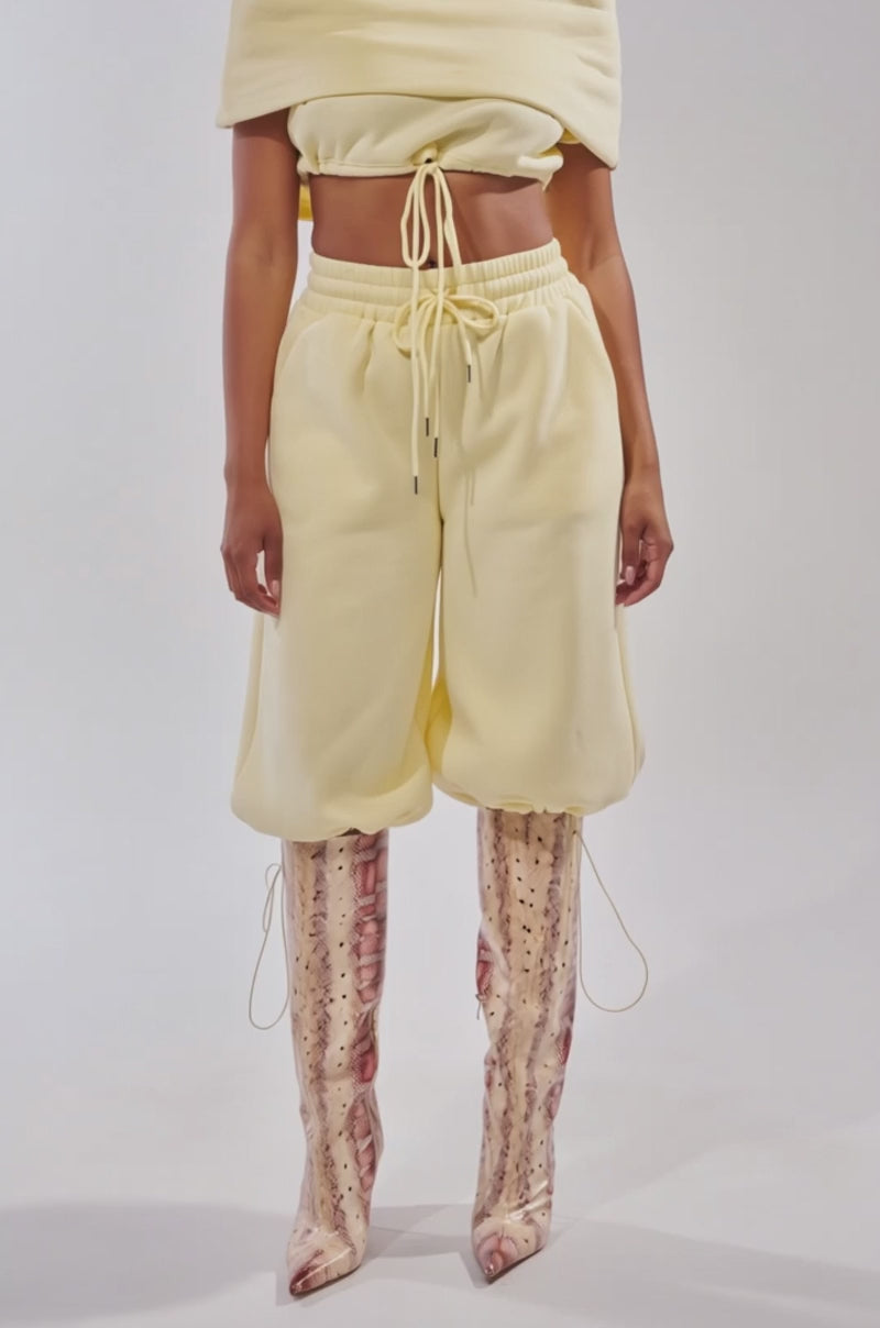 A video of a woman wearing the BABY PLEASE SWEATSHIRT in baby yellow—a cowl-neck cropped style—paired with matching wide-leg pants and knee-high snakeskin boots.