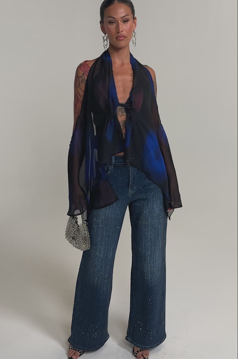 Video of woman modeling the ITS ELECTRIC RHINESTONE WIDE LEG DENIM, featuring high-waisted, wide-leg blue jeans with sparkling rhinestones, paired with black open-toe heels and a sheer black and blue top. 