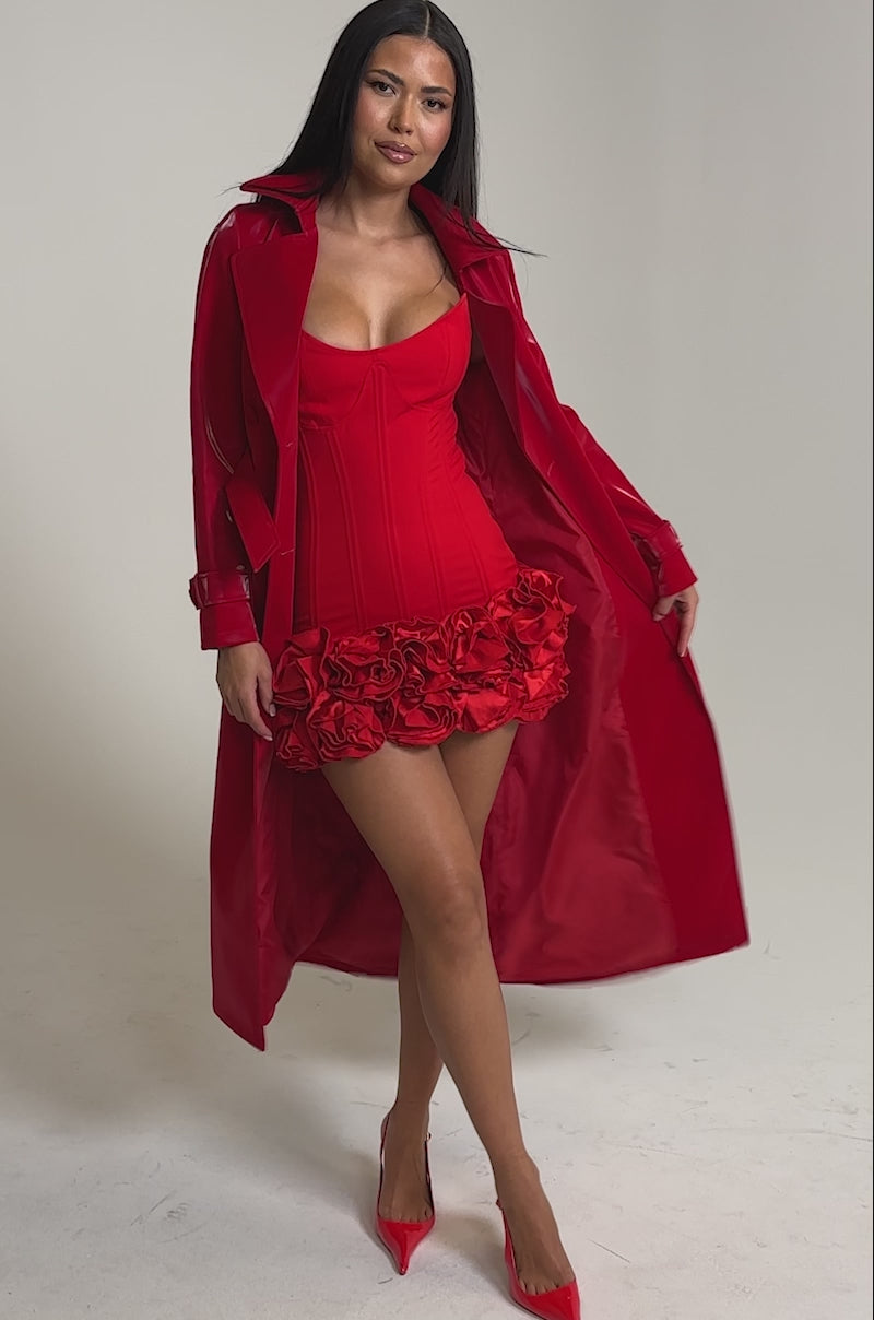 A video of a woman in the DOZEN ROSES LIQUID VELVET TRENCH—a shiny red faux leather coat—styled over a fitted corset dress with rose detailing and red heels, making a striking statement outerwear against a simple background.