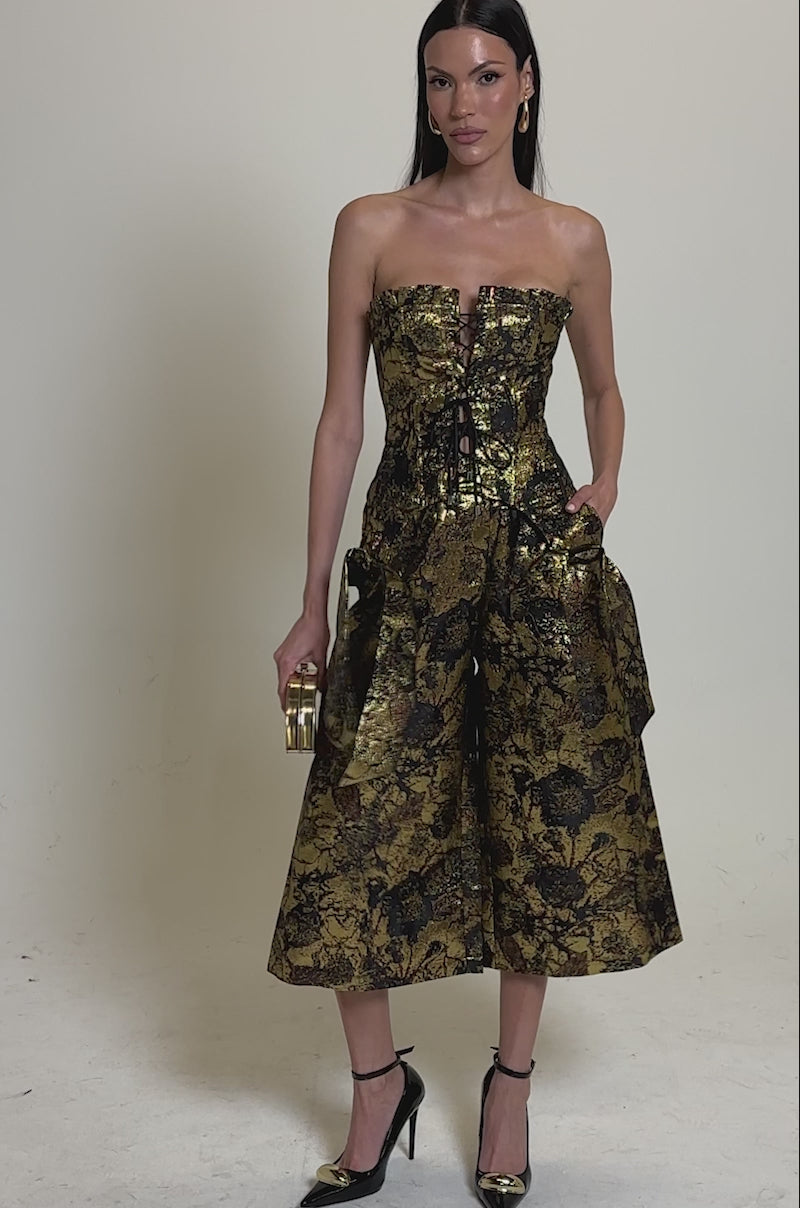 A video of a woman in the LOTUS BROCADE CORSET, a gold-and-black floral corset jumpsuit featuring a lace-up front and flared culotte pants. She accessorizes with a small clutch and black ankle-strap heels.
