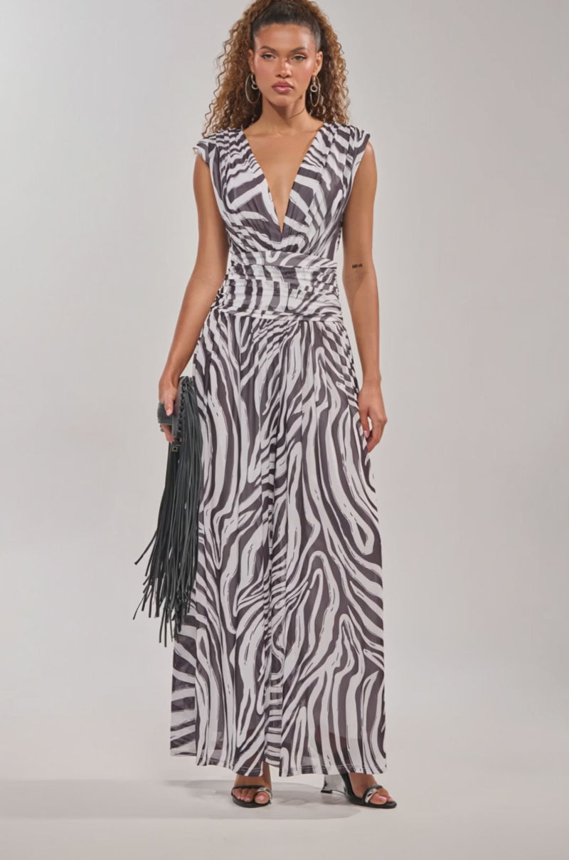 Video of woman modeling the Jungle Fever Zebra Print Jumpsuit, a sleeveless v-neck, floor-length piece with a gathered waist. She holds a black fringed clutch and stands in high-heeled sandals against a plain background.