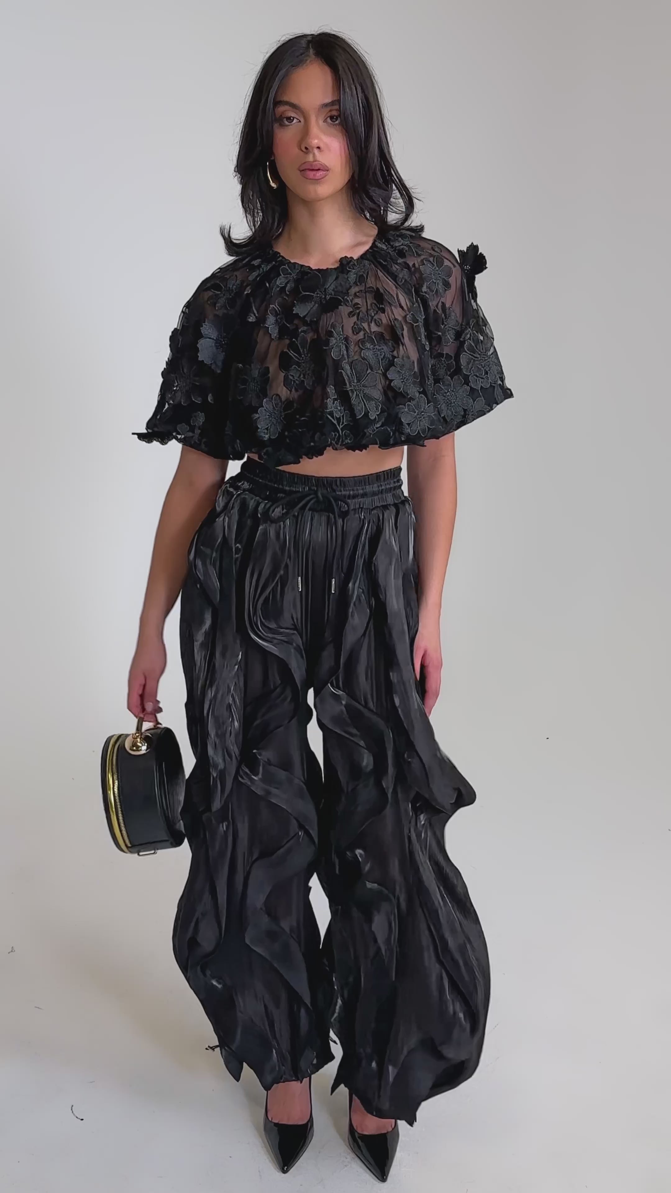 A video of a woman in a sheer, ruffled black crop top and avant-garde street style COOLEST ON THE BLOCK JOGGER pants.