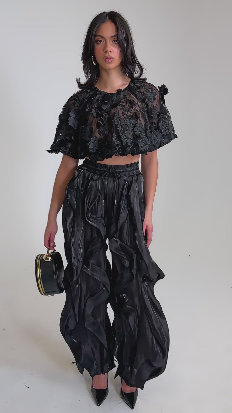 A video of a woman in a sheer, ruffled black crop top and avant-garde street style COOLEST ON THE BLOCK JOGGER pants.