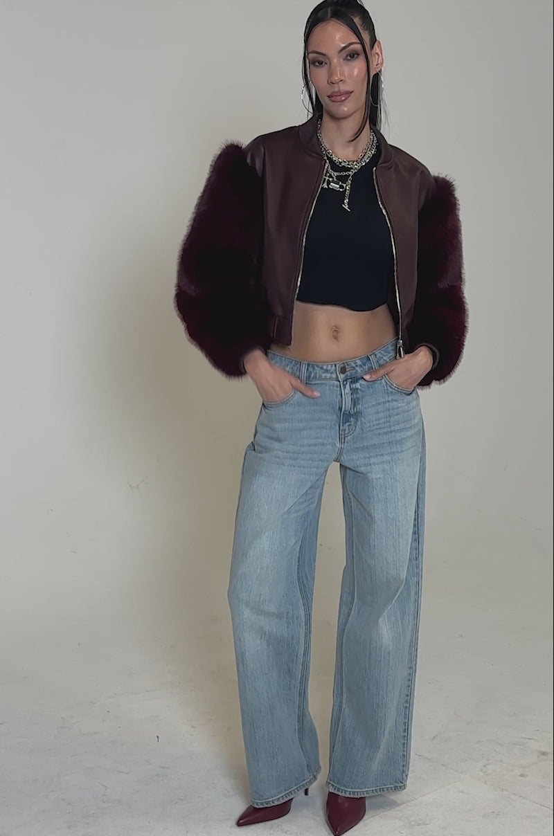 A video of a woman posing in a black cropped top, SO GOOD IT HURTS FUR SLEEVE BOMBER IN BURGUNDY, light blue wide-leg jeans, and maroon heeled boots. Her long dark hair is tied back and she wears layered necklaces against a plain background.