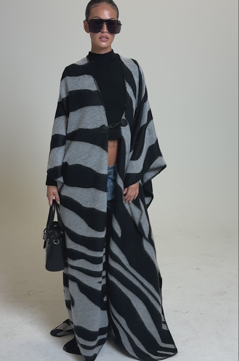 A video of a model wearing oversized sunglasses, a black top, blue jeans, and holding a black handbag, a person stands on a neutral background in the WILD ONE KNIT MAXI PONCHO, featuring a long, flowing zebra pattern.