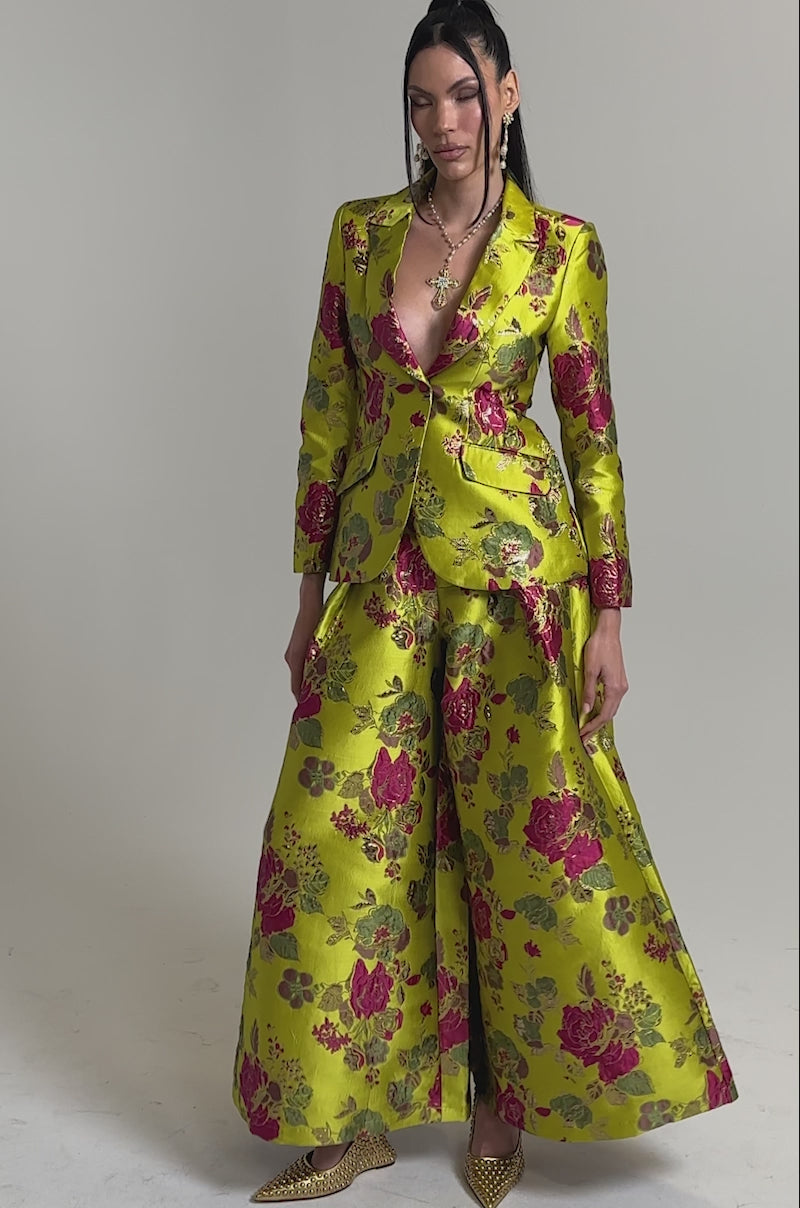 A video of a woman wearing the BUY MYSELF FLOWERS BROCADE BLAZER IN LIME, a vibrant chartreuse fitted blazer with a bold pink and gold floral pattern, paired with wide-leg trousers, gold heels, and layered jewelry against a plain light background.