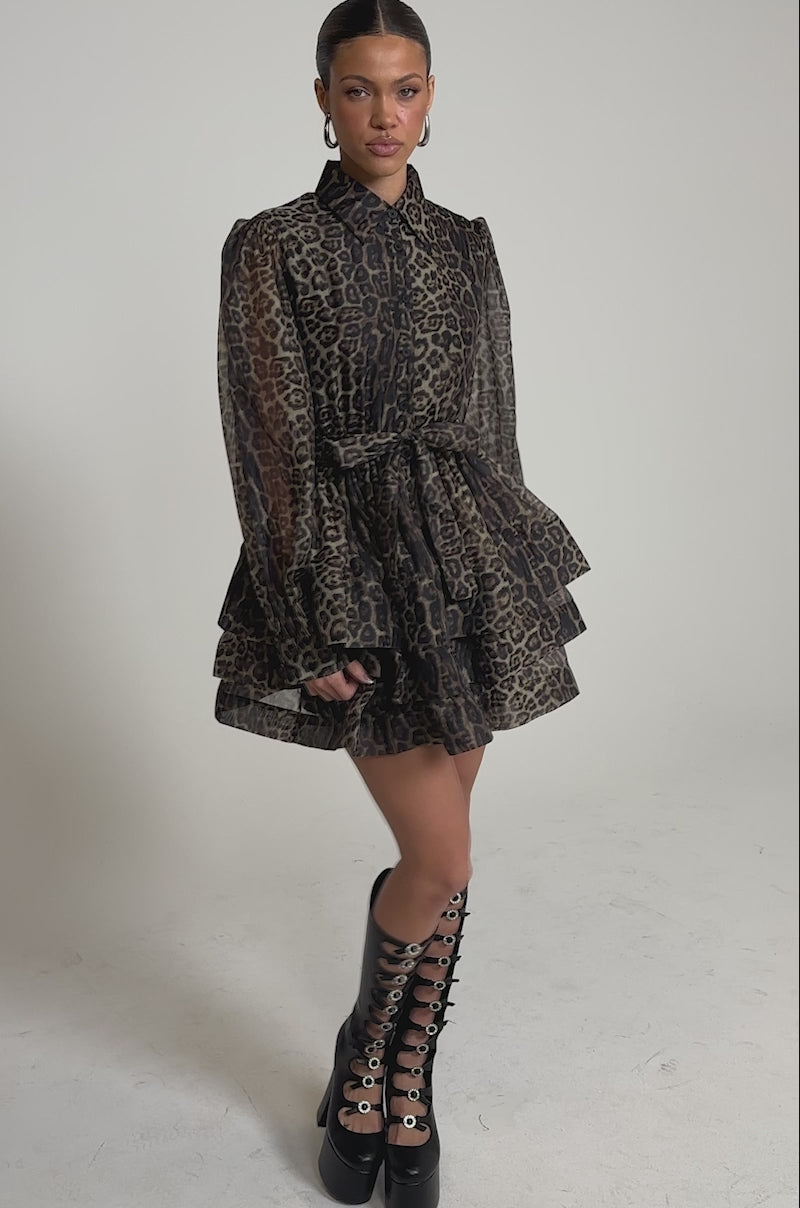 A video of a woman wearing the CHICEST OF CHIC LEOPARD PRINT MINI DRESS—mini, sheer, ruffled, belted at the waist with long sleeves—paired with knee-high black lace-up boots featuring large metal eyelets.