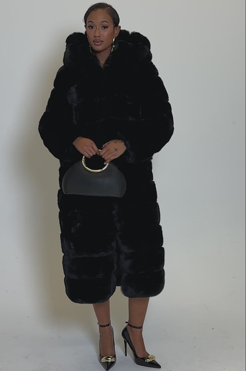 Video of woman modeling the FAUX FUR PANEL HOODED TRENCH IN BLACK over a strapless black dress with gold chains, black heels, gold earrings, and holding a black handbag, a woman poses confidently against a plain light background.