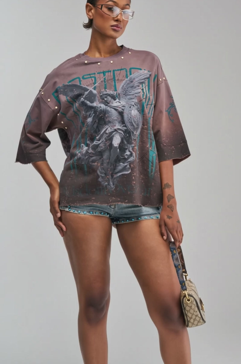 Video of a woman modeling the FIRST ROW WISDOM GRAPHIC TEE—a loose, oversized brown shirt with an angel print.