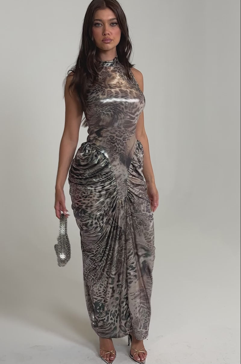 Video of woman modeling the STANDING MY GROUND MOCK NECK MAXI DRESS, a sleeveless, ruched maxi with an abstract animal print. She accessorizes with strappy heels and a sparkling handbag.