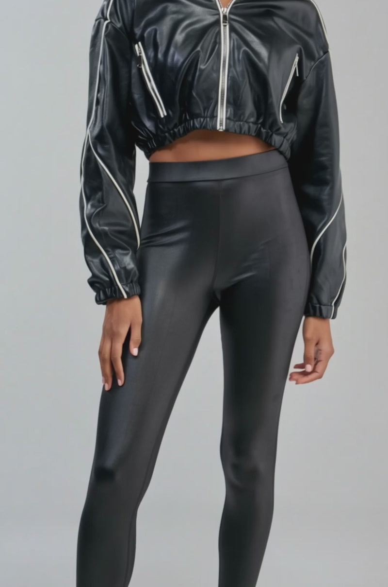 Video of a woman modeling the FAUX LEATHER HOODED BOMBER in glossy black with white piping.